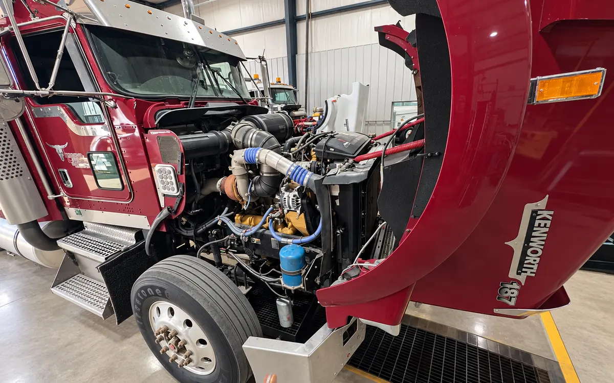 Burgundy Kenworth over the service pit with the hood tilted forward for maintenance